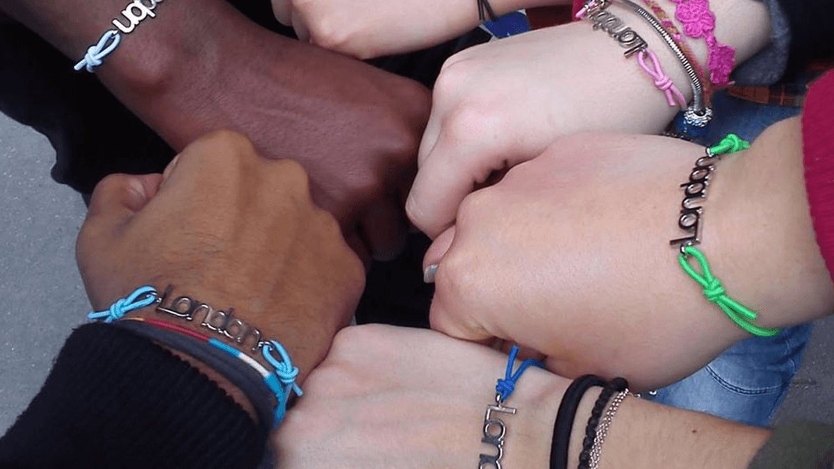 A group of young people from different ethnicities, each with a frniedship bracelt, put their hands into a circle