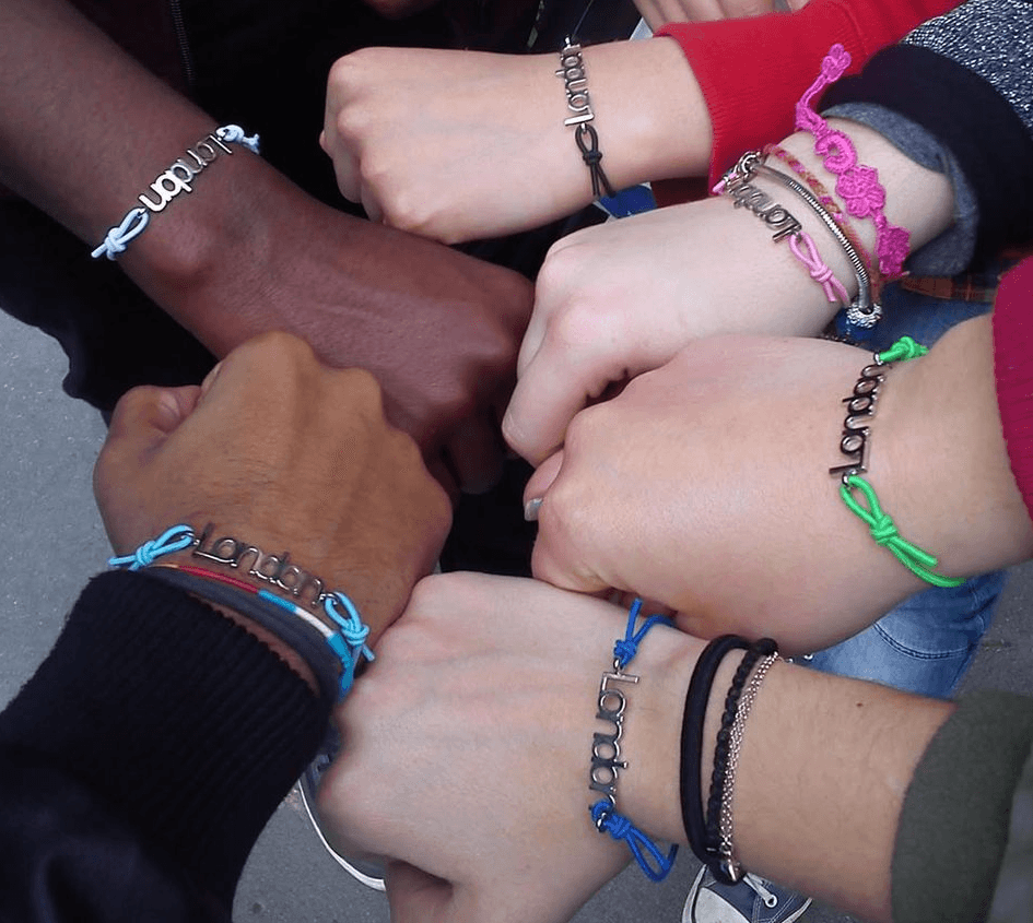 A group of young people from different ethnicities, each with a frniedship bracelt, put their hands into a circle