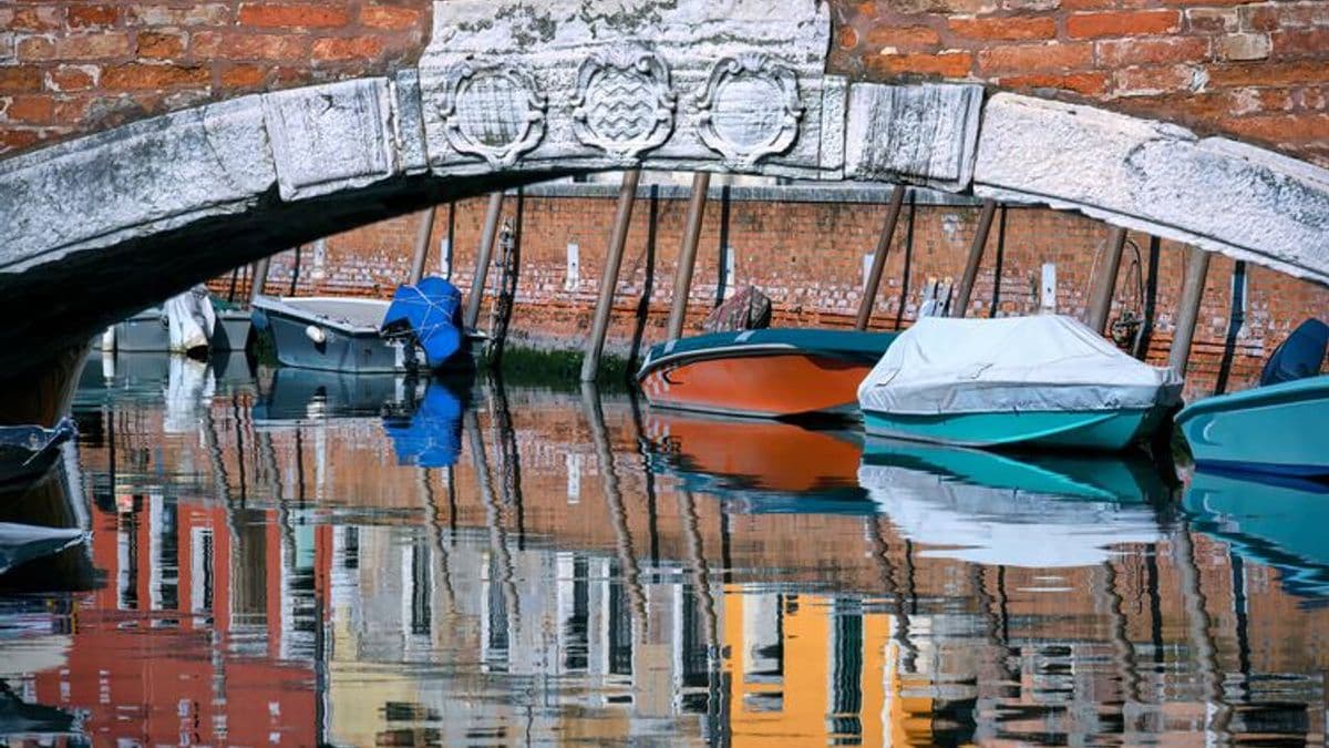 View under a brick bridge of moored boats reflected in the water of a canal, Venice, Italy
