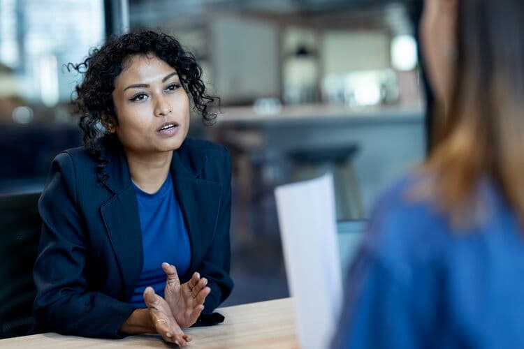 A woman sits in an office environment advising another woman who we only see from the back